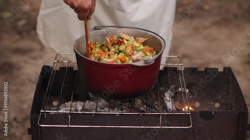 Chef stirring vegetable stew with mushrooms in a large pot on a grill, cooking food over an open fire outdoors.