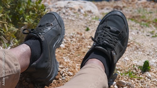 First person view of a man resting during a hike. Personal perspective of a tired hiker resting on a rocky trail after a long trek, with his legs outstretched and feet in black walking shoes crossed