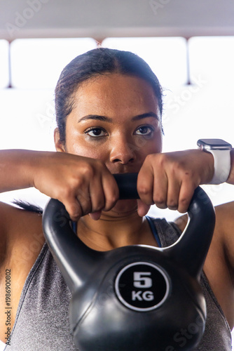 Asian woman holding black 5 KG kettlebell at chest in gym wearing gray tanktop white smartwatch