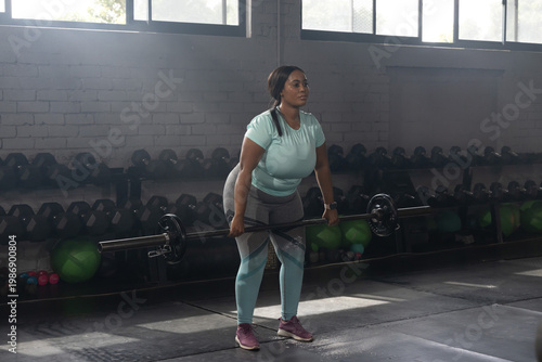 African American woman lifting barbell in gym weight room, wearing teal top and patterned leggings