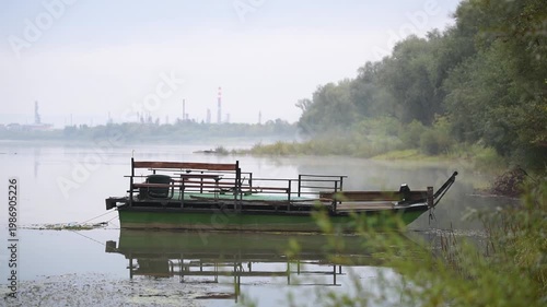 Moored boat floats in river near riverbank on Sava river near Brod, oil refinery in fog in background
