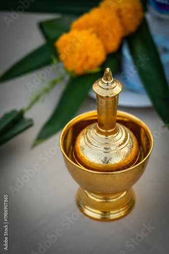Traditional Thai golden brass water pouring container set used for Buddhist merit-making ceremonies with marigold flowers in the background.