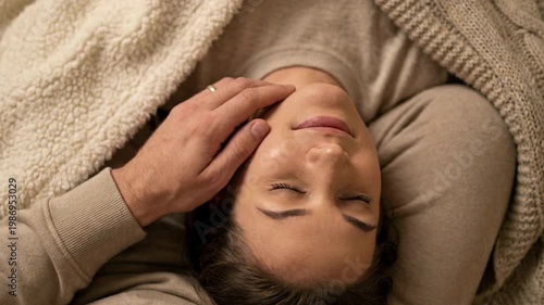 Person resting with closed eyes while partner gently touching face under soft blanket, calm home setting, relaxation, care, wellness routine, comfort, intimacy and peaceful evening moment.