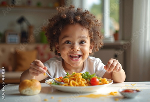A young, curly-haired child with light brown skin sits at a table, smiling broadly while eating pasta