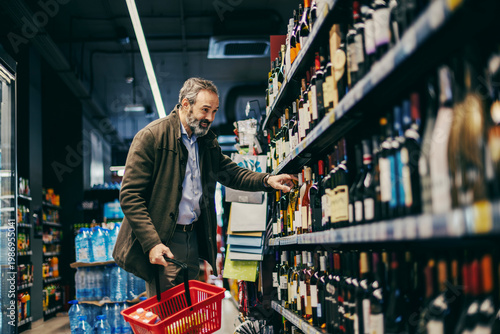 Tableau sur toile Mature man choosing wine bottle in supermarket aisle