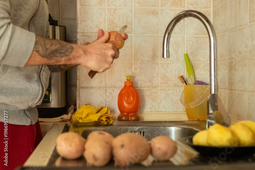 A man, of whom only his arms are framed, cuts the peel of a potato using a knife. The man uses the kitchen sink as a support point
