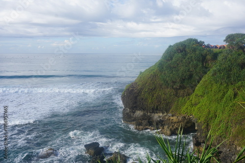 Fotografie Green cliff with dense vegetation overlooking ocean waves crashing against rocky shoreline under partly cloudy sky