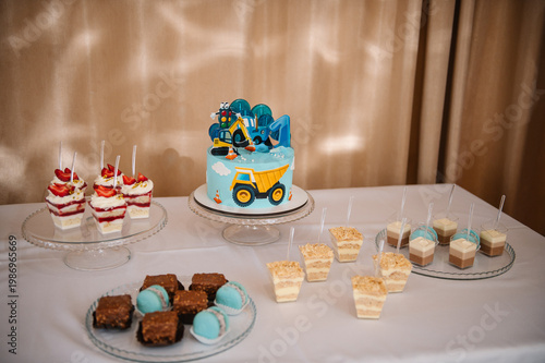 Candy bar for a boy's 1st birthday with a construction themed cake, strawberry trifles, macarons, and mousse desserts. Festive sweet table at a restaurant with blue and yellow treats.