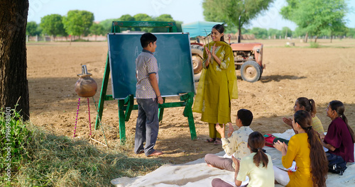 Indian happy young adult gen z female do work talk to villager girl boy sitting on mat rural area. teen male child writing study look board clap hand enjoy desi life open day class field ground place