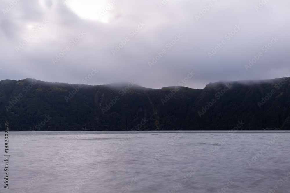 Obraz premium Sete Cidades volcanic lake in San Miguel Island, Azores, Portugal. Calm water reflects cloudy sky. Lush green hills surround the crater lake.