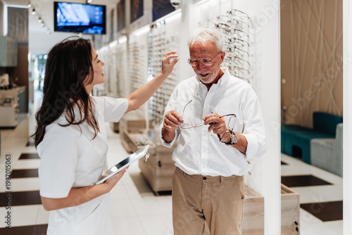 Fotografija An elderly man receives professional help from a female optician while choosing and fitting new glasses in a bright, modern optical shop