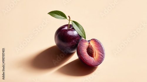Fresh purple plum with green leaf beside a halved plum showing the inner flesh and seed on a smooth beige background