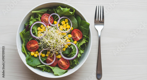 A white bowl filled with a fresh mix of greens, tomatoes, onions, corn, and sprouts sits next to a fork on a light wooden table