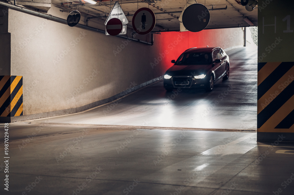 Naklejka premium Car exits underground parking garage in urban area during evening hour with signs visible and concrete surface illuminated by headlights