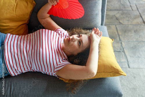 Woman cooling herself with red hand fan while lying on sofa at home