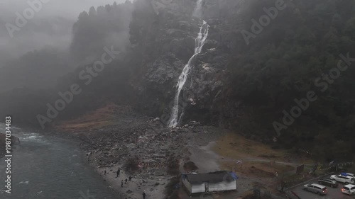 Aerial drone footage captures a tall waterfall cascading down rugged rocky cliffs into a misty mountain valley with turquoise river waters and small tourist structures and vehicles parked below surrou
