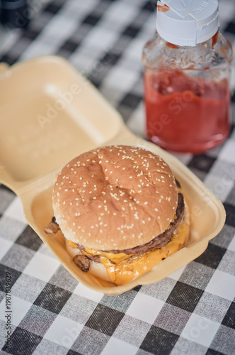 Close up of a cheeseburger in a takeaway container set on a black and white checkered tablecloth, with warm golden bun tones and melted cheese. Classic uk cafe culture.