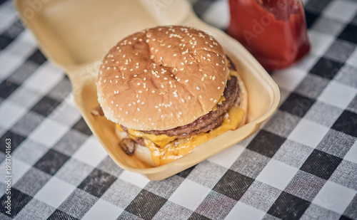 Close up of a cheeseburger in a takeaway container set on a black and white checkered tablecloth, with warm golden bun tones and melted cheese. Classic uk cafe culture.