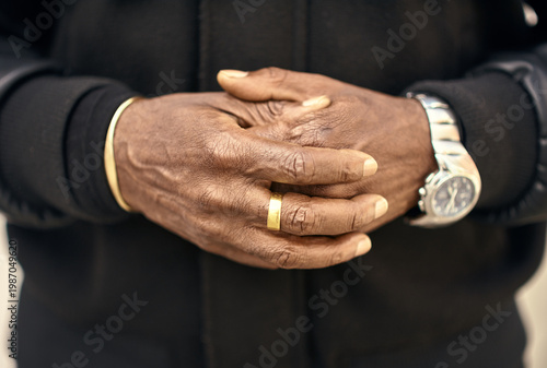 Close up of clasped hands with rich brown skin tones, gold ring and silver watch adding subtle highlights against a deep black garment. Confident hand gesture.