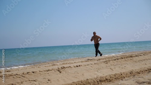 Shirtless man jogging along sandy beach, showcasing dynamic movement and energy, with gentle waves lapping at shore under clear blue sky, capturing the essence of outdoor fitness and vitality