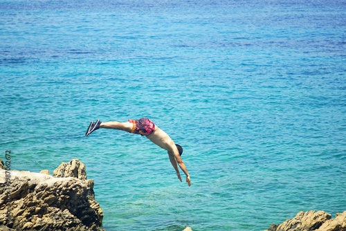Man diving headfirst from rugged coastal rocks into turquoise sea under sunlight. Reveals cliff jumping culture, adrenaline seeking tourism, embodied freedom practice, and experiential travel trend.