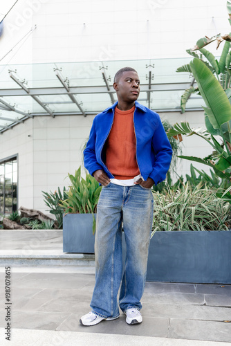 A young African man stands in a greenhouse surrounded by various plants. He wears a blue jacket over an orange sweater and jeans.