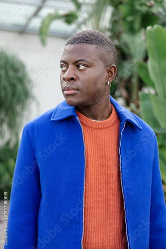 A young African man stands in a lush green environment. He has short black hair and wears a blue jacket over an orange sweater.