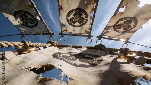 Low angle shot of tattered traditional Japanese samurai banners with family crests against a bright blue sky. Historical war flags with ropes and metal rings at a cultural festival