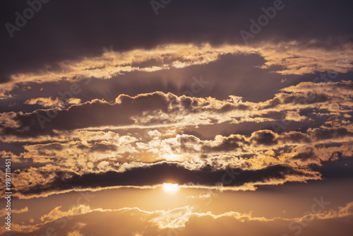 Golden sun setting behind dark storm clouds, dramatic rays breaking through creating ominous glow in moody landscape