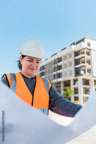 A young Hispanic woman in a hard hat and orange vest examines blueprints at a construction site. A modern building is visible in the background.