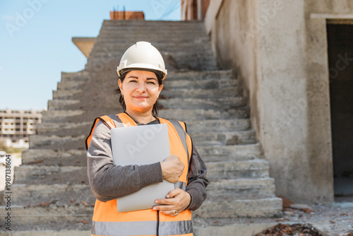 Bilde på lerret woman stands on construction stairs wearing a hard hat and safety vest, holding a laptop