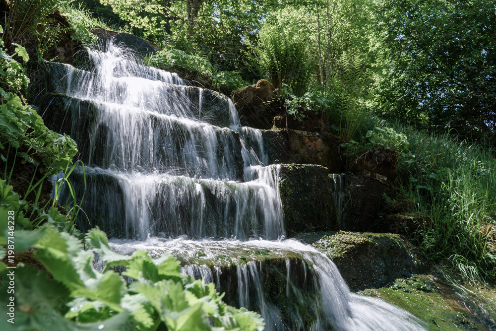 Obraz premium Cascading forest waterfall flowing smoothly over stone steps in long exposure with silky water motion and soft misty atmosphere in summer sunlight