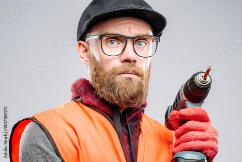 A worker with a power drill stands in a studio, wearing an orange vest, glasses, red gloves, and a black cap while looking seriously at the camera