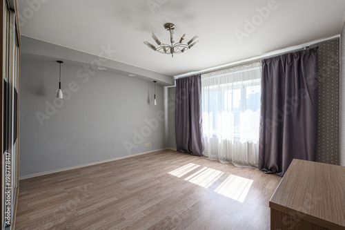 A spacious, light room with light grey walls, wooden flooring, a chandelier, pendant lights, large window with sheer and dark curtains, partial furniture.