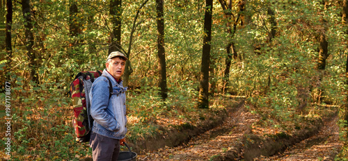 Man with a large backpack and a bucket walking on a dirt road through an autumn forest. Side view of a hiker exploring nature during golden hour. Wide panoramic shot with copy space