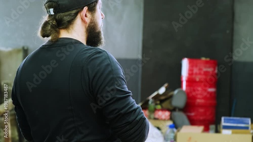 Young bearded man mechanic stands in garage backside view. Skilled technician in black uniform waits car to repair at service station slow motion