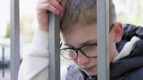 A teenage boy stands behind a metal fence, looking upset and isolated. The scene conveys sadness and bullying at school, highlighting emotional struggle and vulnerability. 