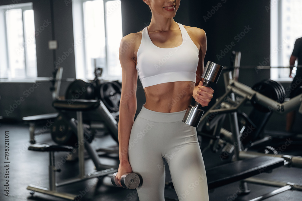 Fototapeta premium Female fitness enthusiast in white sports bra and gray leggings holds dumbbells in a gym, with exercise equipment and large windows visible in the background