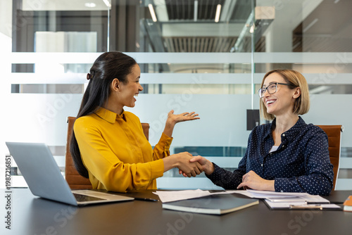 Photos Two businesswomen are smiling and shaking hands across an office desk with a lap