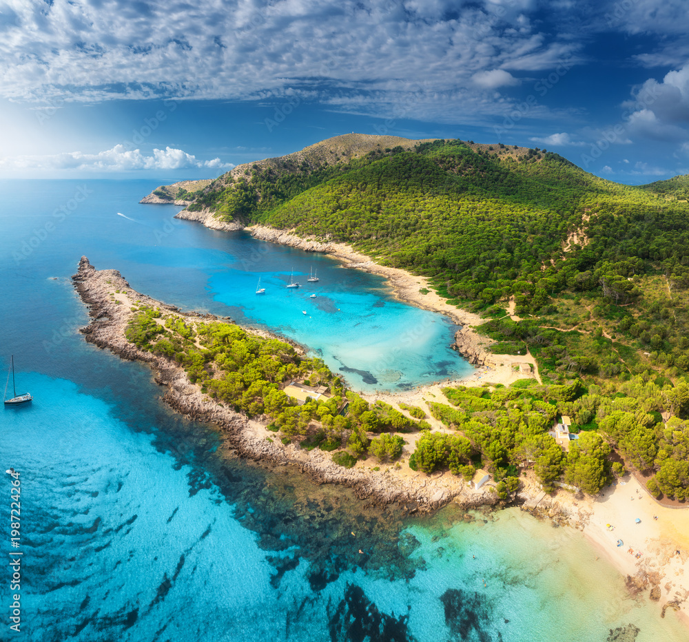 Obraz premium Aerial view of Cala Agulla beach, turquoise water, white sand, lush greenery, yachts and boats anchored in the picturesque bay of Mallorca, Balearic Islands, Spain, Summer travel. Top drone view