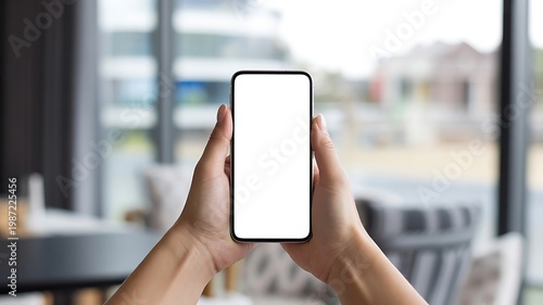 A person holding a smartphone with a blank white screen in their hands indoors