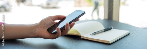 A womans hand holding a smartphone next to an open notebook and pen on a table