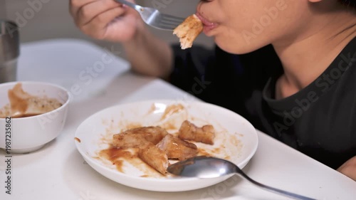 A boy eating pork steak with a fork from a white plate at home. The image reflects homemade food, mealtime, childhood lifestyle, appetite, and everyday family dining.