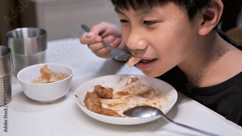 A boy smiling while eating pork steak with a fork at home. The image reflects homemade food, enjoyable mealtime, childhood lifestyle, appetite, and everyday family dining.