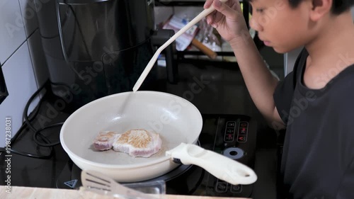 Close-up of a 10 year old child cooking pork steak in a pan on a stovetop. The image reflects learning, cooking practice, and life skills development in a home kitchen.