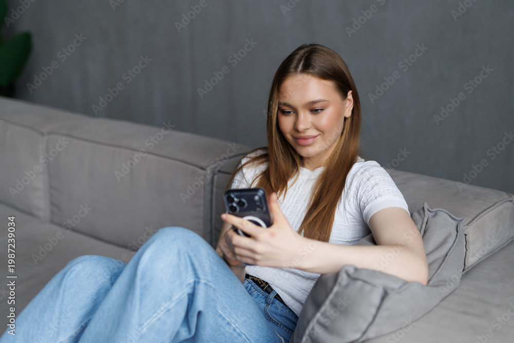 Obraz premium Happy young woman using mobile phone and smiling while sitting on sofa at home