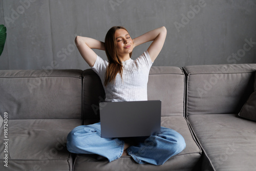 Young woman is stretching her arms while sitting on a sofa working remotely on her laptop in her cozy living room