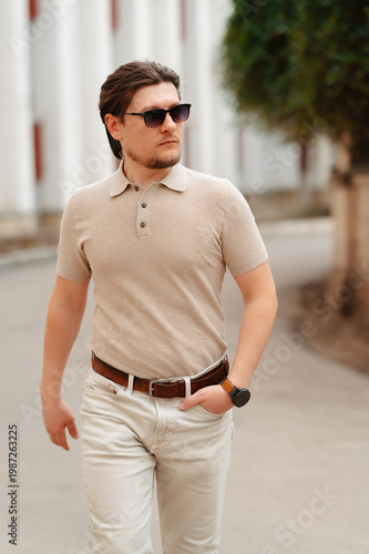 Caucasian Man In Beige Polo, Striding Along Classical Colonnade Under Warm Sunlight, Sunglasses Reflecting
