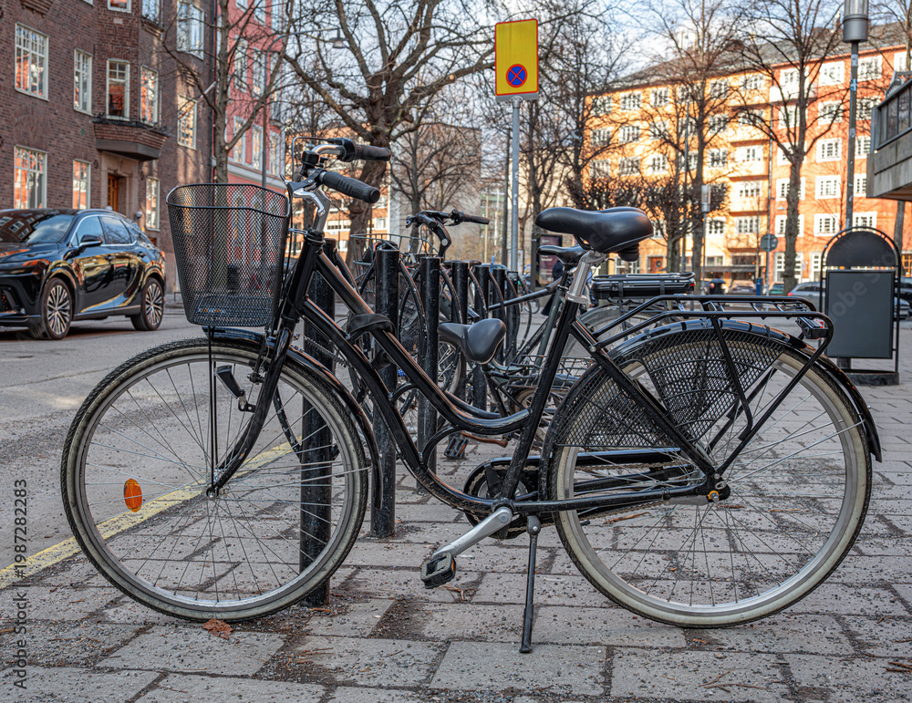 Fototapeta premium A classic black city bicycle with a front basket is parked on a stone-paved sidewalk in a quiet urban neighborhood.
