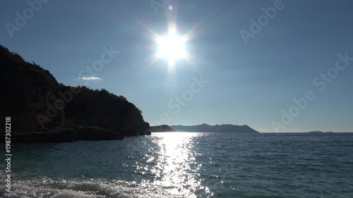 Kas, Turkey - 24 of February 2026: Bright sun with rays reflecting on the sea and dark rocky cliffs under blue sky
